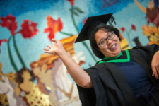 Female graduate wearing glasses, smiling and waving in front of a colourful mural